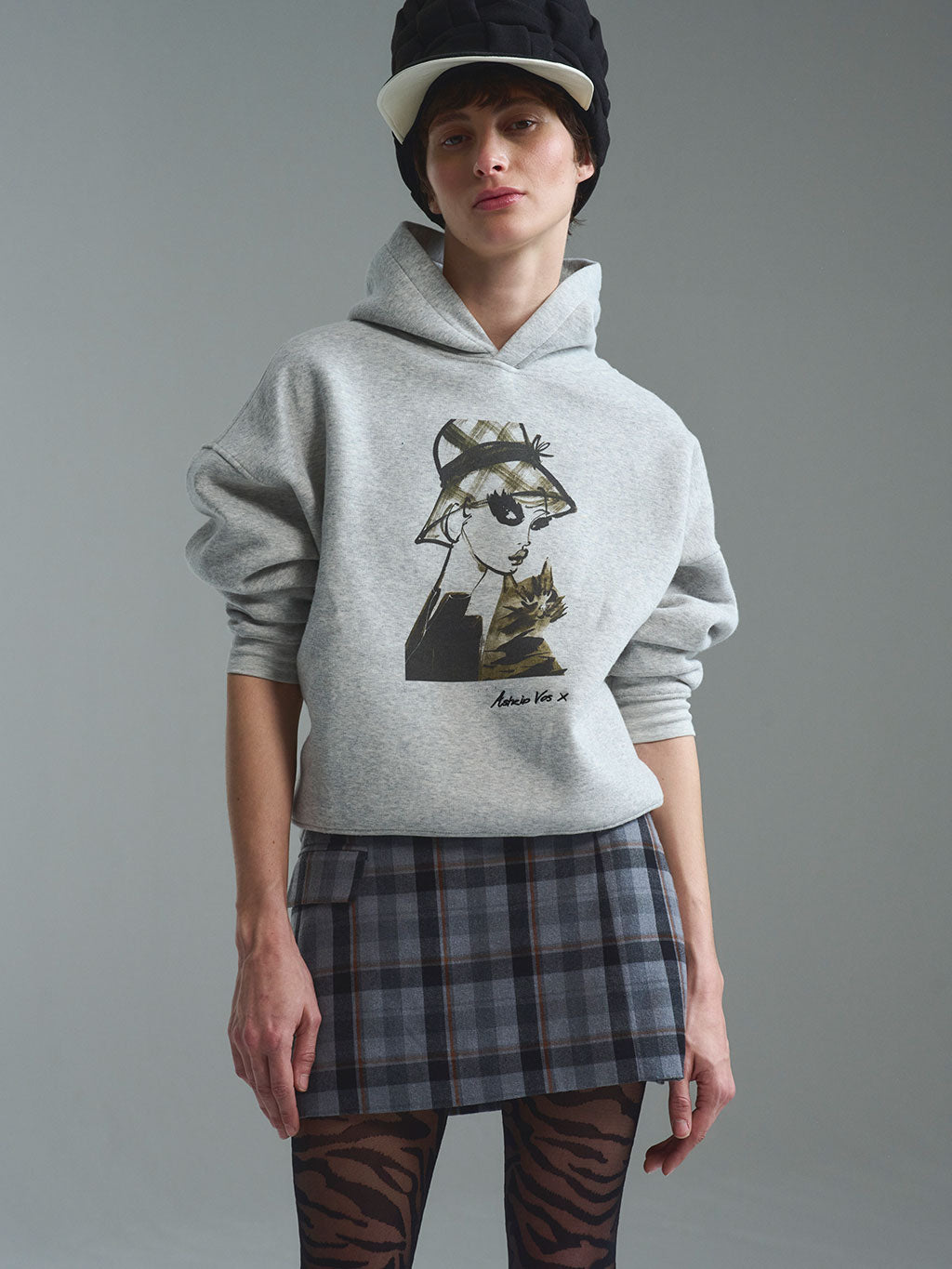 Exklusiver grauer Damen-Kapuzenpullover aus der limitierten RAILIN x Astrid Vos X Art Edition. Der hochwertige, aufwändige Art Print zeigt die ausdrucksstarke Modeillustration Innocent glance mit einer Frau im Karo-Hut und einer Katze als tragbares Kunstwerk.
